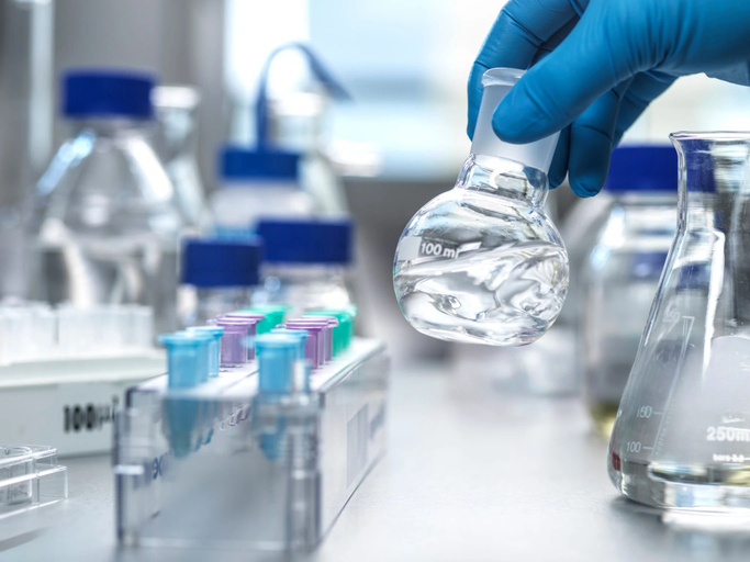 A hand wearing blue gloves lifts up some lab equipment in a chemical formulation and manufacturing laboratory